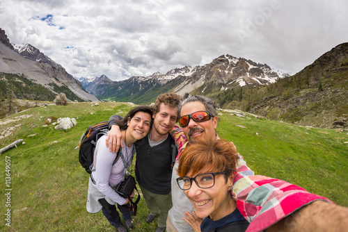Four young people taking selfie on the Alps with snowcapped mountain range and dramatic sky in background. Scenic fisheye distortion. Concept of traveling people and nature beauty exploration.