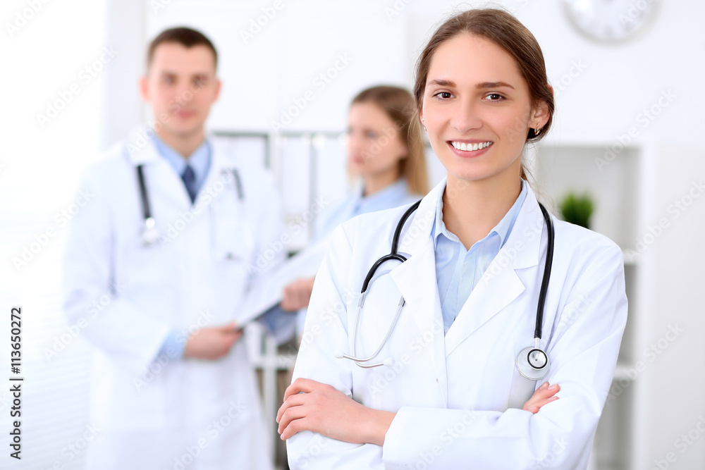 Young beautiful female doctor smiling  on the background with patient and his doctor in hospital. High level and quality medical service concept.