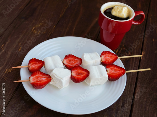 A cup of coffee and marshmallows with fresh strawberries on skewers on a wooden background.