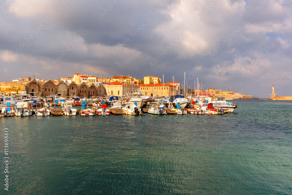 Chania Arsenals, the Venetian shipyards, and fishing boats in old harbour of Chania with Lighthouse in cloudy summer morning, Crete, Greece