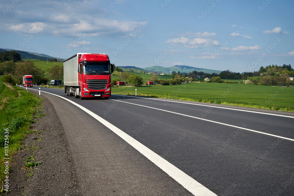 Red trucks arriving on the asphalt road winding through the green fields. Villages and forested mountains in the background. Sunny spring day with blue skies and white clouds.