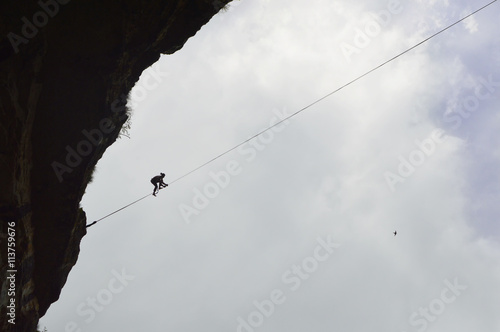Silhouette of vertical cliffs, young highline walker on a tightrope high in the cloudy sky