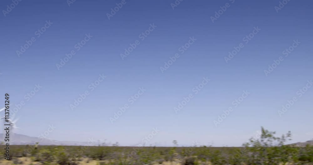 Pan from a mountain view to wind turbines generating power in heat haze on scrubby desert land at a wind farm near the Tehachapi Mountains in California. Blue sky with a few clouds. Pan from a mountain view to wind turbines generating power in heat haze on scrubby desert land at a wind farm near the Tehachapi Mountains in California. Blue sky with a few clouds.
