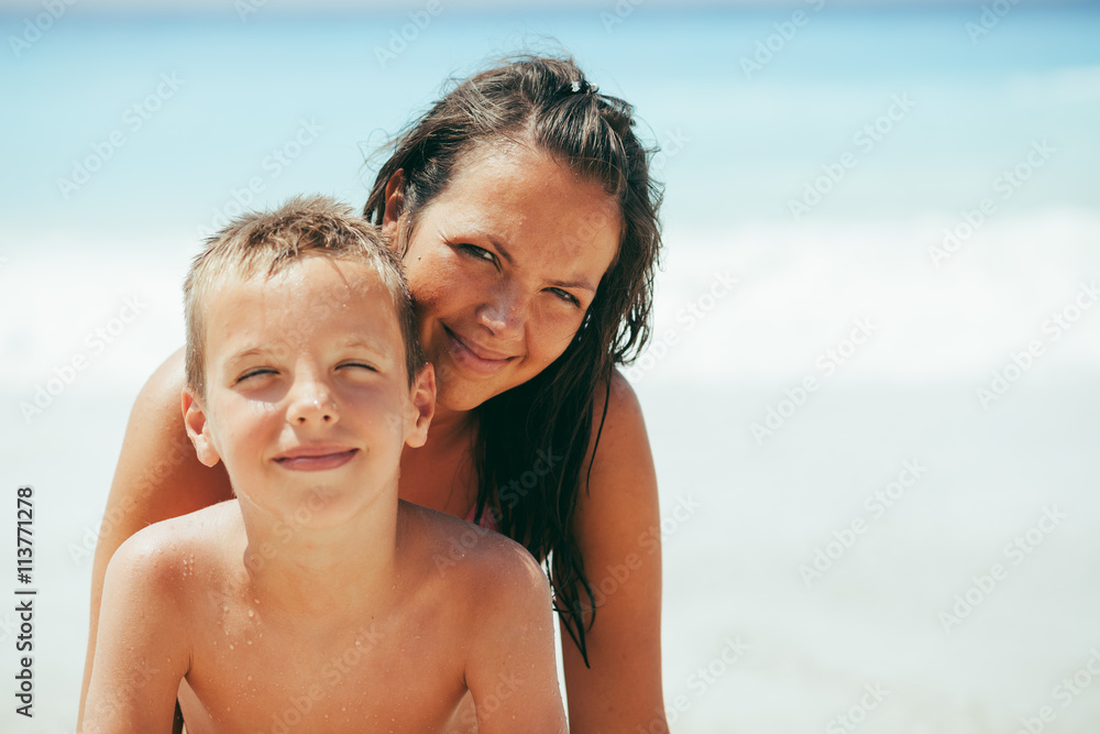Young mother and her son looking at camera, smiling and posing on beautiful sunny tropical beach. Sea in the background.