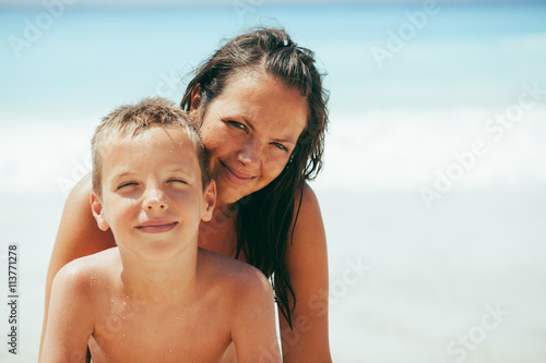 Young mother and her son looking at camera, smiling and posing on beautiful sunny tropical beach. Sea in the background. © Dusko