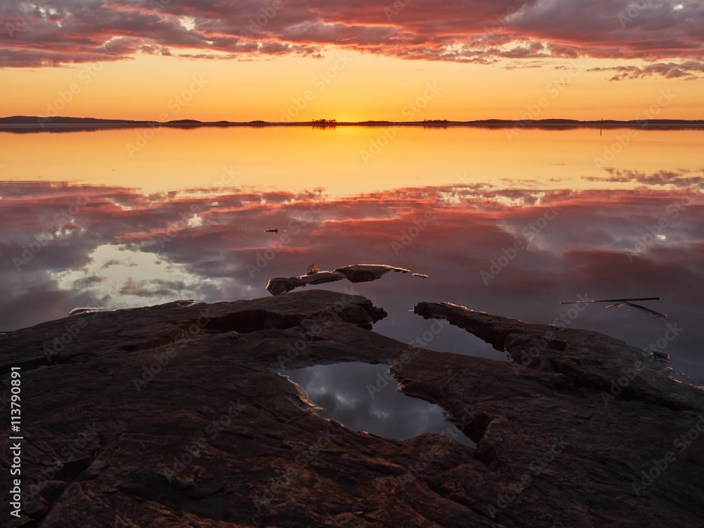 Sunset with vibrant red color in the clouds and dark rocks on the shore. Dramatic reflection of the sunset in the still water of a lake in Finland.
