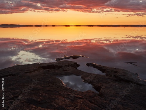 Sunset with vibrant red color in the clouds and dark rocks on the shore. Dramatic reflection of the sunset in the still water of a lake in Finland.