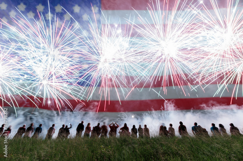 Group of people enjoying fireworks show in a carnival or holiday. People in silhouette and blurred American flag in background.