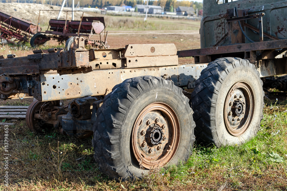 The old broken truck with two big lowered wheels against the village with agricultural machinery The old broken truck with two big lowered wheels against the village with agricultural machinery