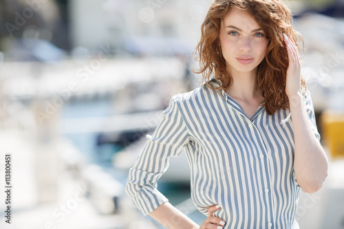Stylish young woman with gray eyes,curly red hair and sweet smile,dressed in white pants and a bright striped shirt,walks on the pier next to the yacht club and the sea, yachts and boats