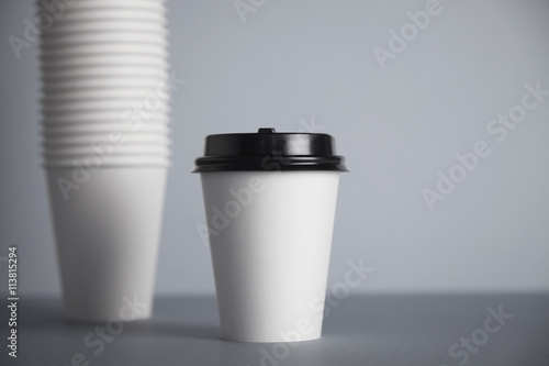 One focused take away white paper cup with black cap presented in front of unfocused group of other cups folded one in other in big bunch, isolated on simple gray background
