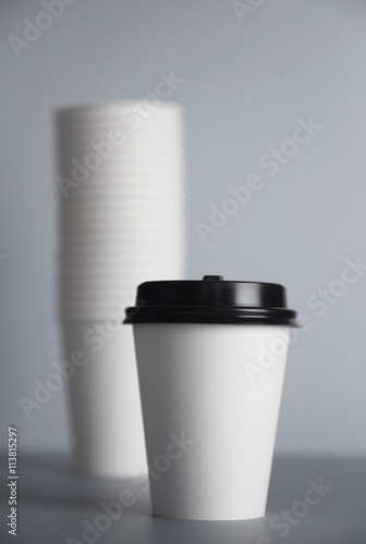 One focused take away white paper cup with black cap presented in front of unfocused group of other cups folded one in other in big bunch, isolated on simple gray background, close view