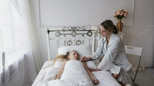 Woman doctor with a stethoscope auscultating to the child lying in the bed.Doctor or nurse examines the girl in the bed stethoscope.The doctor in a white room with a stethoscope.Woman doctor with a