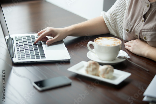 Hands of beautiful young woman sitting at table with cup of coffee and cakes and working on laptop. Attractive model using computer in cafe or office interior. Close-up