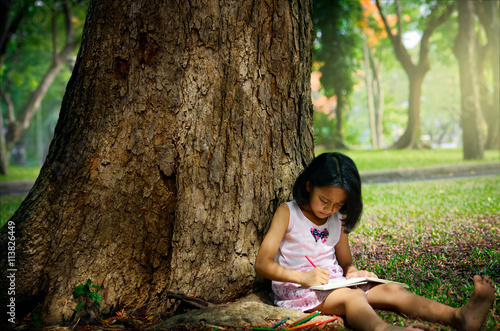 Little girl sitting under a big tree and drawing picture. her it is verry cheerful photo