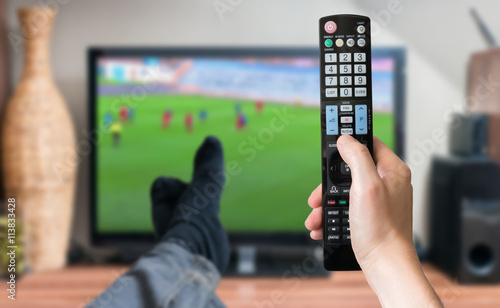 Man is relaxing with legs on table and is watching football match on tv with remote controller.