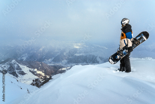 Snowboarder standing on cliff in snowy weather in the mountains.
