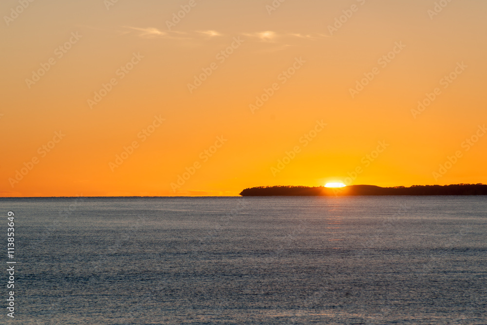 Bright orange sky above flat ocean at sunrise with low island shimmering on horizon.