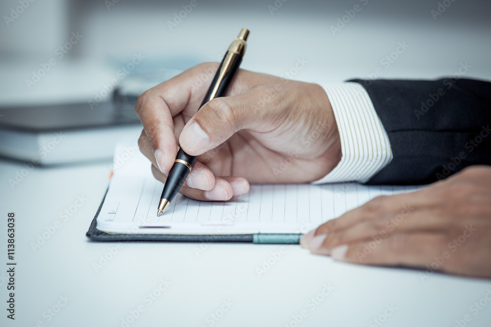Businessman writing on notebook with pen in the office in blue c Businessman writing on notebook with pen in the office in blue c