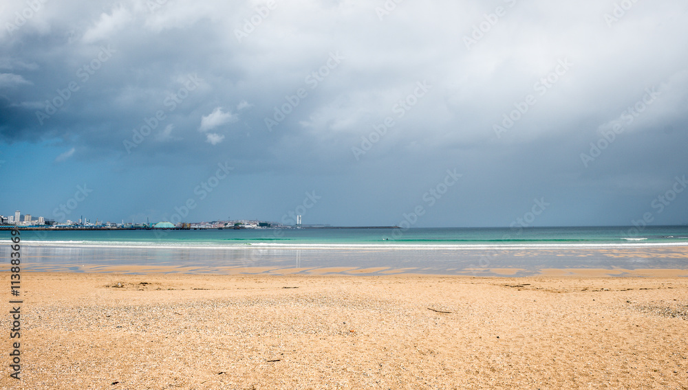 Tropical sandy beach with storm clouds and city in background.
