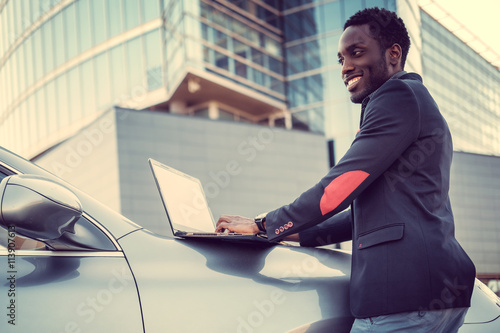 Stylish african male using laptop near his car.