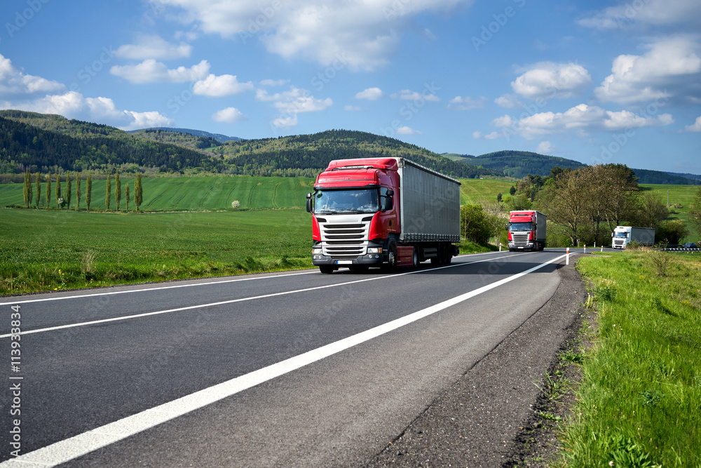 Three trucks driving on asphalt road between green fields in the countryside. Wooded mountains in the background. Sunny day with blue skies and white clouds.