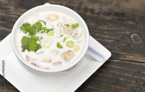 Thai soup in a white dish on a wooden table. Spicy coconut milk soup with shrimp and greens. 