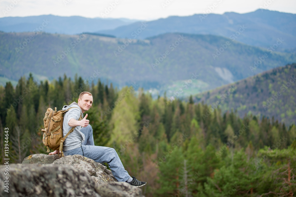 Smiling tourist male with a backpack sitting on the edge of a rock and showing thumb up gesture of good class, with an astonishing blurred view on forest valley and hills