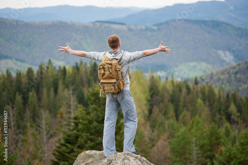 Obraz na płótnie Male climber with brown backpack standing on the top of a rock with back to the camera, his arms out to the sides and enjoying picturesque view of on the green forest and nice mountains w salonie