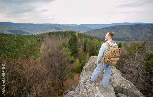 Courageous man with a backpack standing on the edge of a rock and looking into the distance on the green forest and nice mountains © anatoliy_gleb