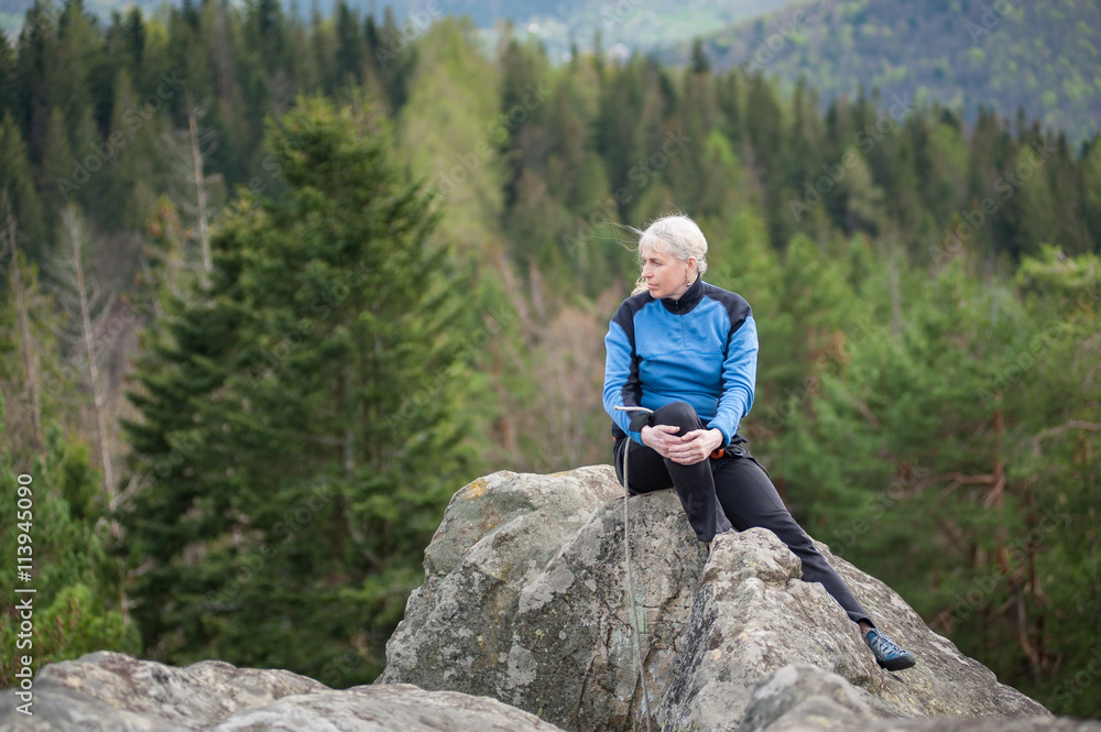 Naklejka premium Athletic old female in a blue jacket with climbing equipment sitting on the top of the rock on the blurred background of forest valley.