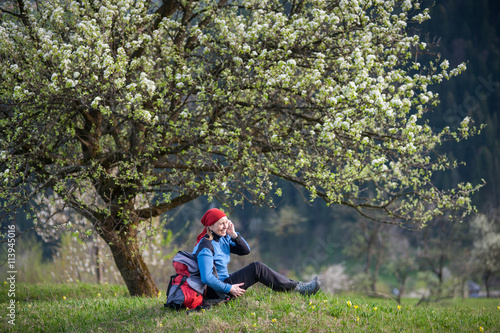 Woman with a backpack enjoy under blooming tree on top of hill with yellow wildflowers, green grass and talking on the mobile phone with forest valleys as blurred background.