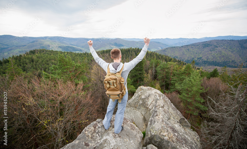 Man traveler with a backpack standing on mountain top, with back to the camera with hands up, against picturesque view of on the green forest and nice mountains. View from above Man traveler with a backpack standing on mountain top, with back to the camera with hands up, against picturesque view of on the green forest and nice mountains. View from above