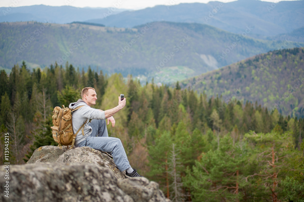 Happy man with a backpack sitting on the edge of a rock and makes selfie on the his phone with forest and mountains in blurred background