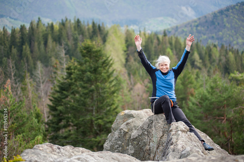 Happy woman in a blue jacket with hands up and with climbing equipment sitting on the top of the rock on the blurred background of forest valley.