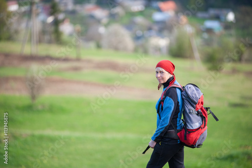 Traveler woman in a red scarf on her head and blue jacket with a backpack looking at the camera, blurred greenery background. Close-up