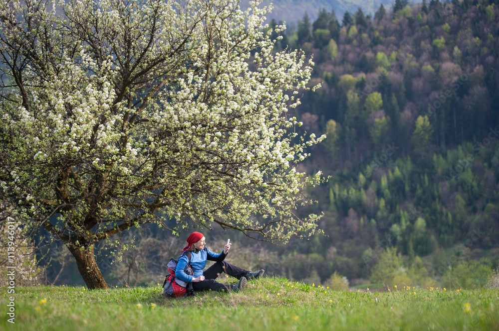 Smiling woman with a backpack sit under blooming tree on top of hill with yellow wildflowers, green grass and makes selfie with forest valleys as blurred background. Smiling woman with a backpack sit under blooming tree on top of hill with yellow wildflowers, green grass and makes selfie with forest valleys as blurred background.