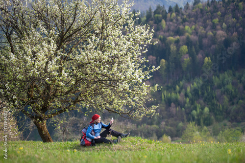 Smiling woman with a backpack sit under blooming tree on top of hill with yellow wildflowers, green grass and makes selfie with forest valleys as blurred background.