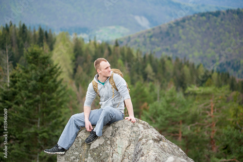 Rock climber with a brown backpack sitting on the peak of rock and looking into the distance on the blurred background of forest valley and hills. Spring nature.