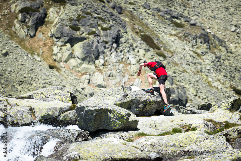 man with backpack running and jumping over stones on high mountain trail with waterfall