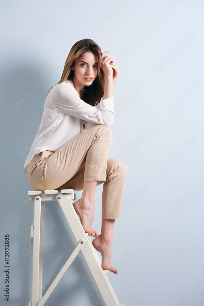 Beautiful young woman sitting on a wooden ladder on blue wall background Beautiful young woman sitting on a wooden ladder on blue wall background