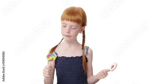 Little  redhead girl is holding two lollypops in both hands, looking at one and the other, smiling, trying one of them, isolated on white background