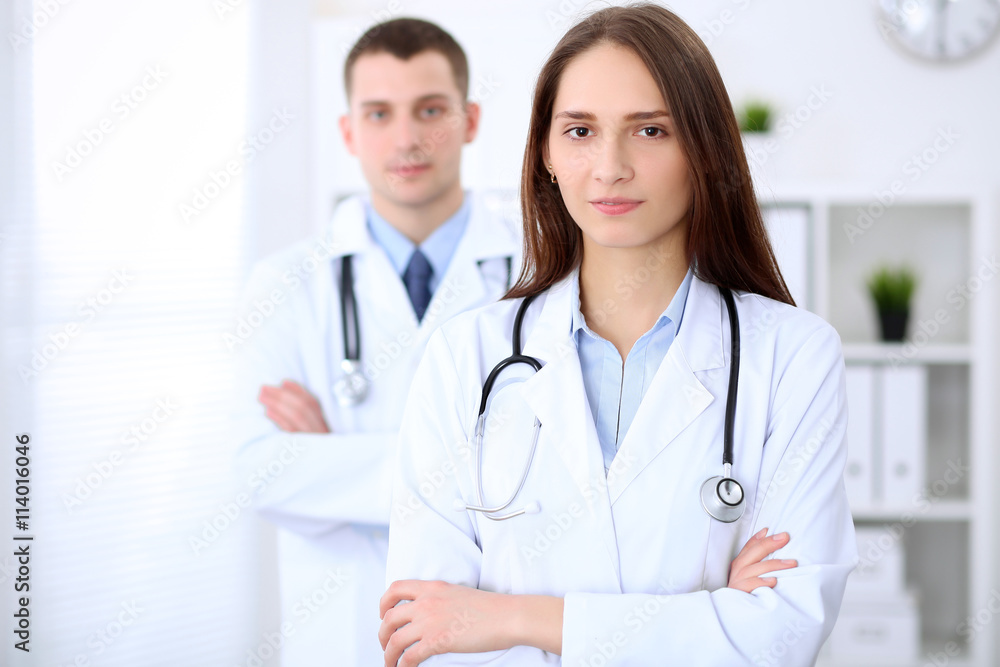 Young beautiful female doctor smiling on the background with male doctor in hospital in hospital. High level and quality medical service concept. Young beautiful female doctor smiling on the background with male doctor in hospital in hospital. High level and quality medical service concept.