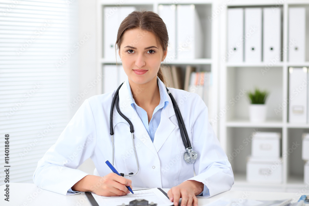 Young brunette female doctor sitting at the table and working at hospital office. Health care, insurance and help concept. Physician ready to examine patient Young brunette female doctor sitting at the table and working at hospital office. Health care, insurance and help concept. Physician ready to examine patient