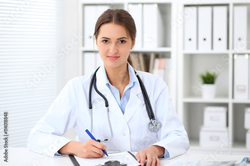 Young brunette female doctor sitting at the table and working at hospital office. Health care, insurance and help concept. Physician ready to examine patient