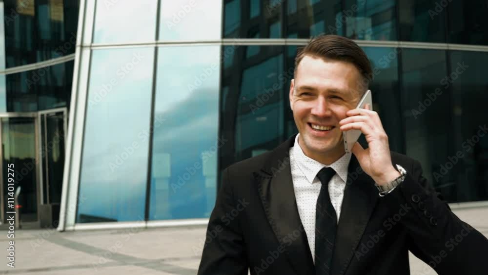 Young European business-man in black suit and tie smiling and talking by smartphone. Modern business-centre building at the background. Teal and Orange style. Close-up 4k flycam shot. Young European business-man in black suit and tie smiling and talking by smartphone. Modern business-centre building at the background. Teal and Orange style. Close-up 4k flycam shot.