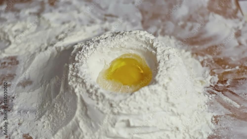 Close up pouring raw egg into well of flour on wooden table Close up pouring raw egg into well of flour on wooden table