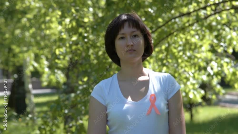 Woman pinning orange ribbon on white t-shirt, trees in background. Leukemia, melanoma, multiple sclerosis, Self Injury Awareness Day Woman pinning orange ribbon on white t-shirt, trees in background. Leukemia, melanoma, multiple sclerosis, Self Injury Awareness Day