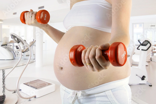 Frau schwanger mit Babybauch trainiert mit Gewichten und Hanteln im Fitness Studio photo