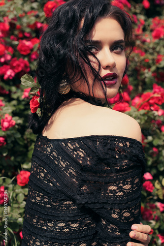 fashion outdoor photo of gorgeous sensual woman with dark hair and bright makeup, in elegant black lace dress posing beside roses at summer garden 